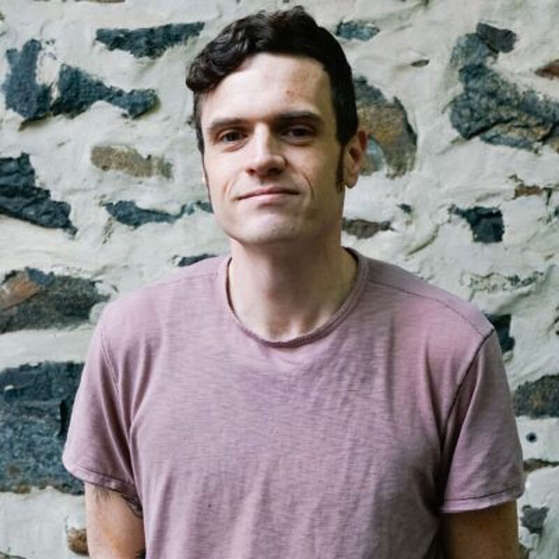 The image shows a man with short, dark curly hair and fair skin, wearing a light purple t-shirt. He is standing in front of a stone wall with varying sizes and colors of rocks. The lighting is soft, highlighting his features and the texture of the wall. He appears to be in his late 20s or early 30s.